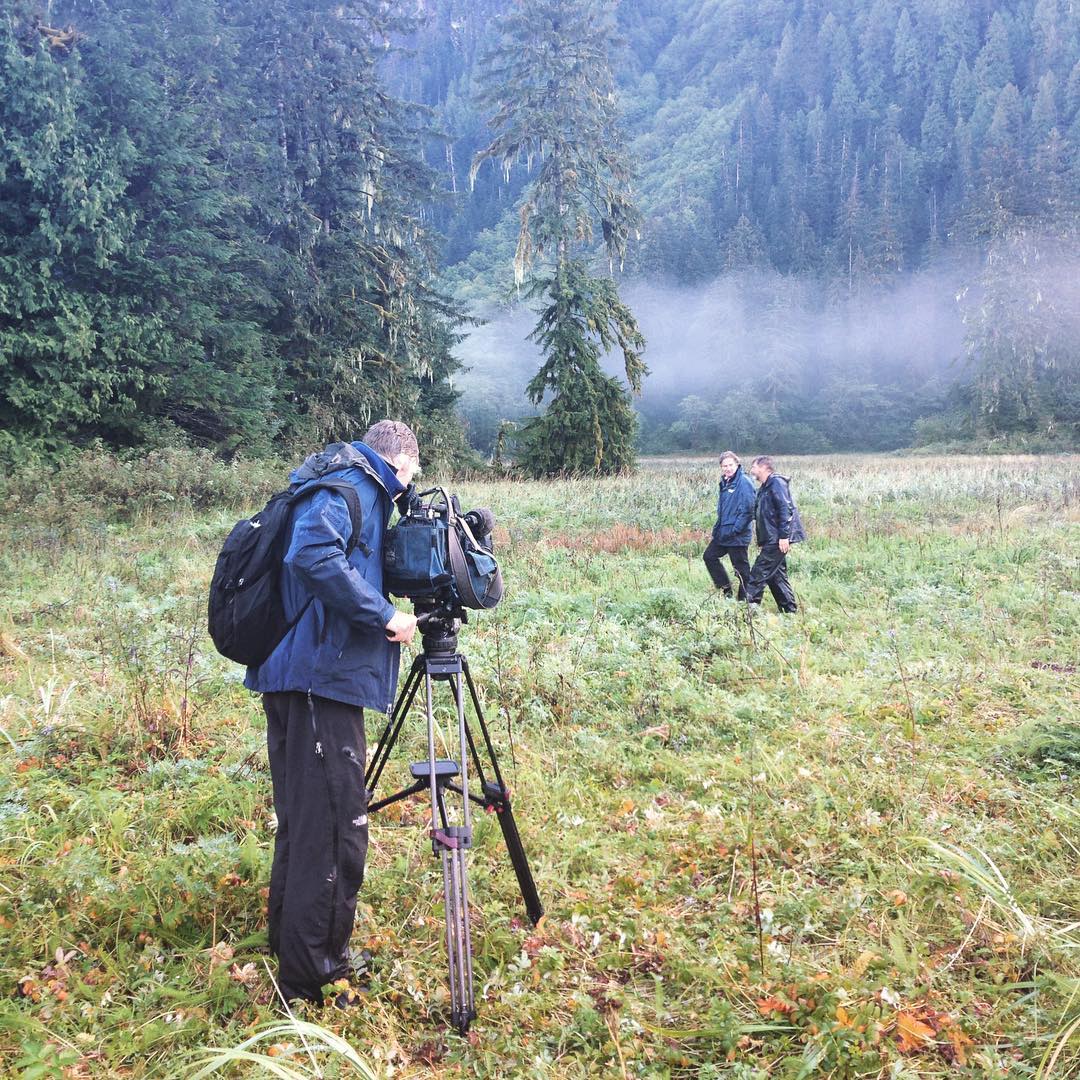A camera person stands behind his camera on a tripod in a clearing in the forest while Raincoast staff Brian Falconer faces the camera