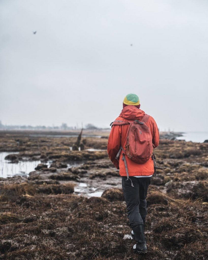 Someone in an orange jacket walking out along a rocky shore on a grey day.