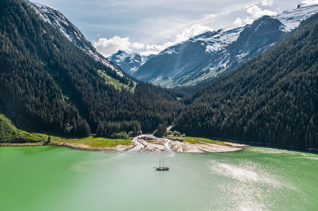 A sail boat on green-tinged water cradles between mountains covered with trees and snow.