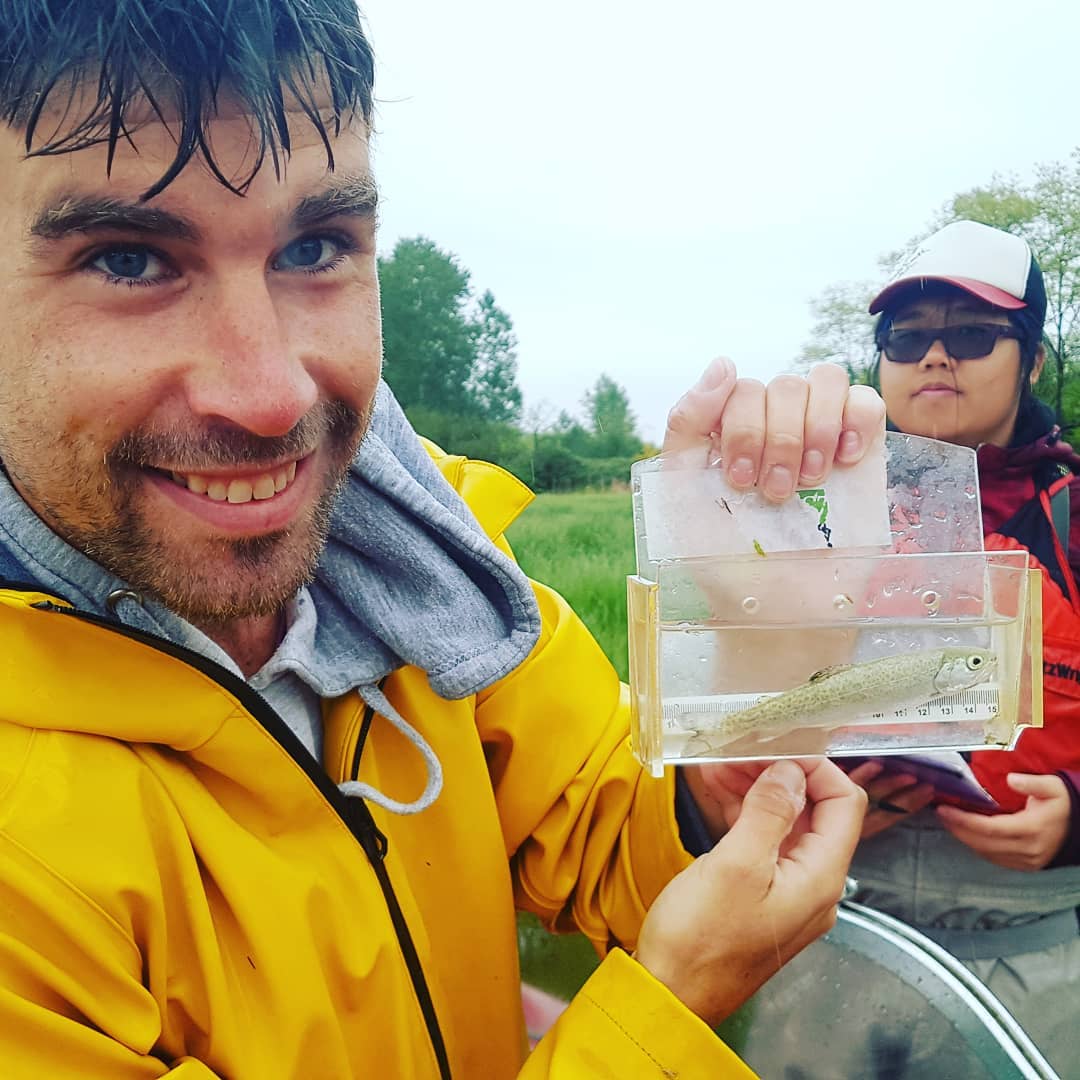 Raincoast staff scientist in yellow rain jacket holds up a plastic sampler bag filled with water and a small fish inside it.