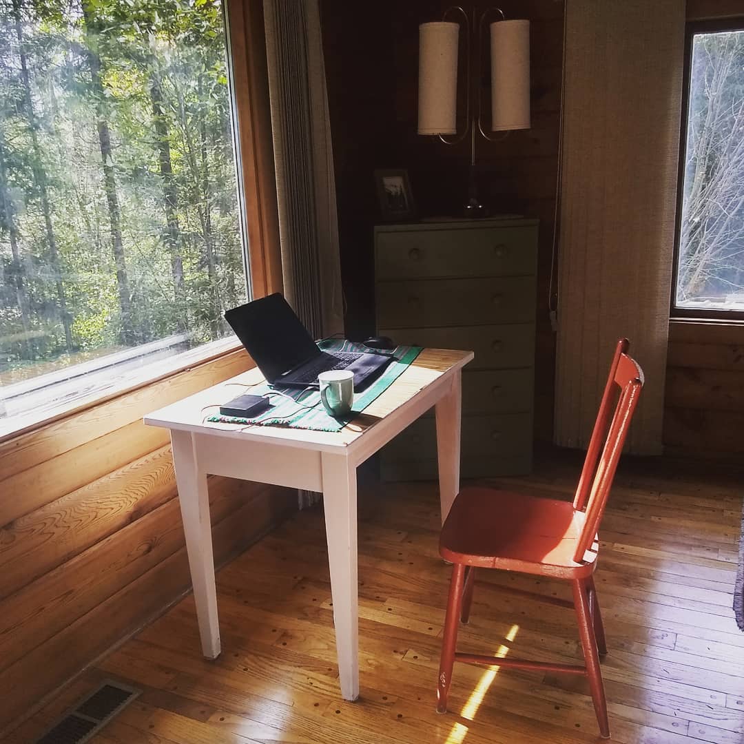 Desk with a laptop computer on it and a red chair facing a window out of a wooden cabin looking out into the forest