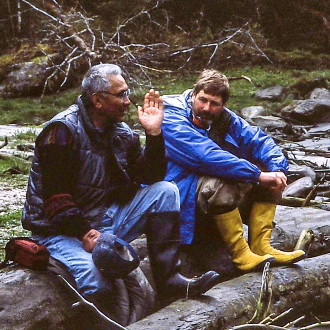 Two men sit on rocks by the water in deep discussion