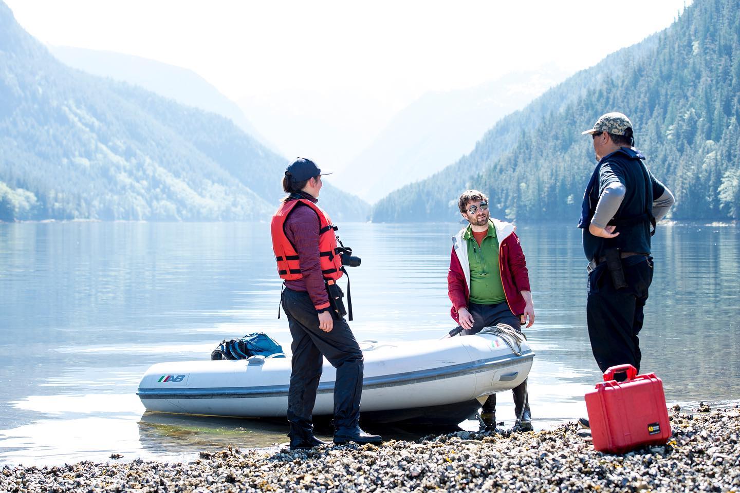 Three Raincoast researchers in life jackets and boots stand on a pebbled shore around a white dinghy speaking to each other while the ocean looms quietly in the background in pale blue, cut by two mountains forming a vee.