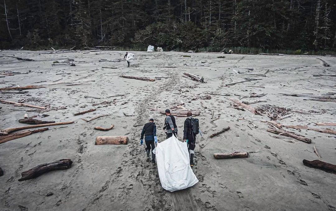 Three people seen from the air pulling garbage in a white bag along sand.