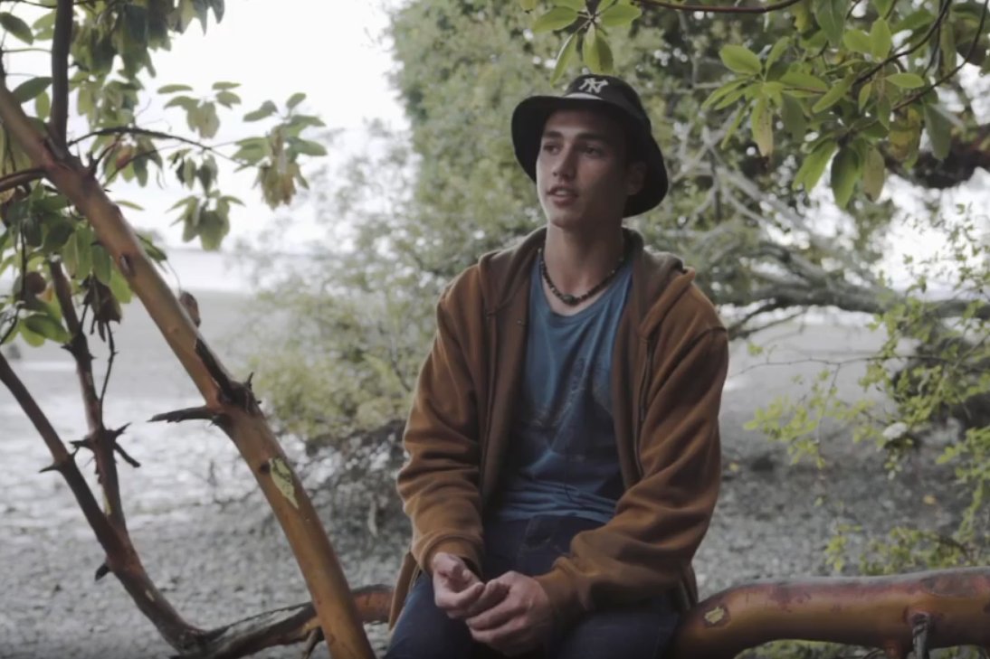 Salish Sea Youth Steward Ryder Wise seated on an arbutus branch by the ocean in a speaking pose