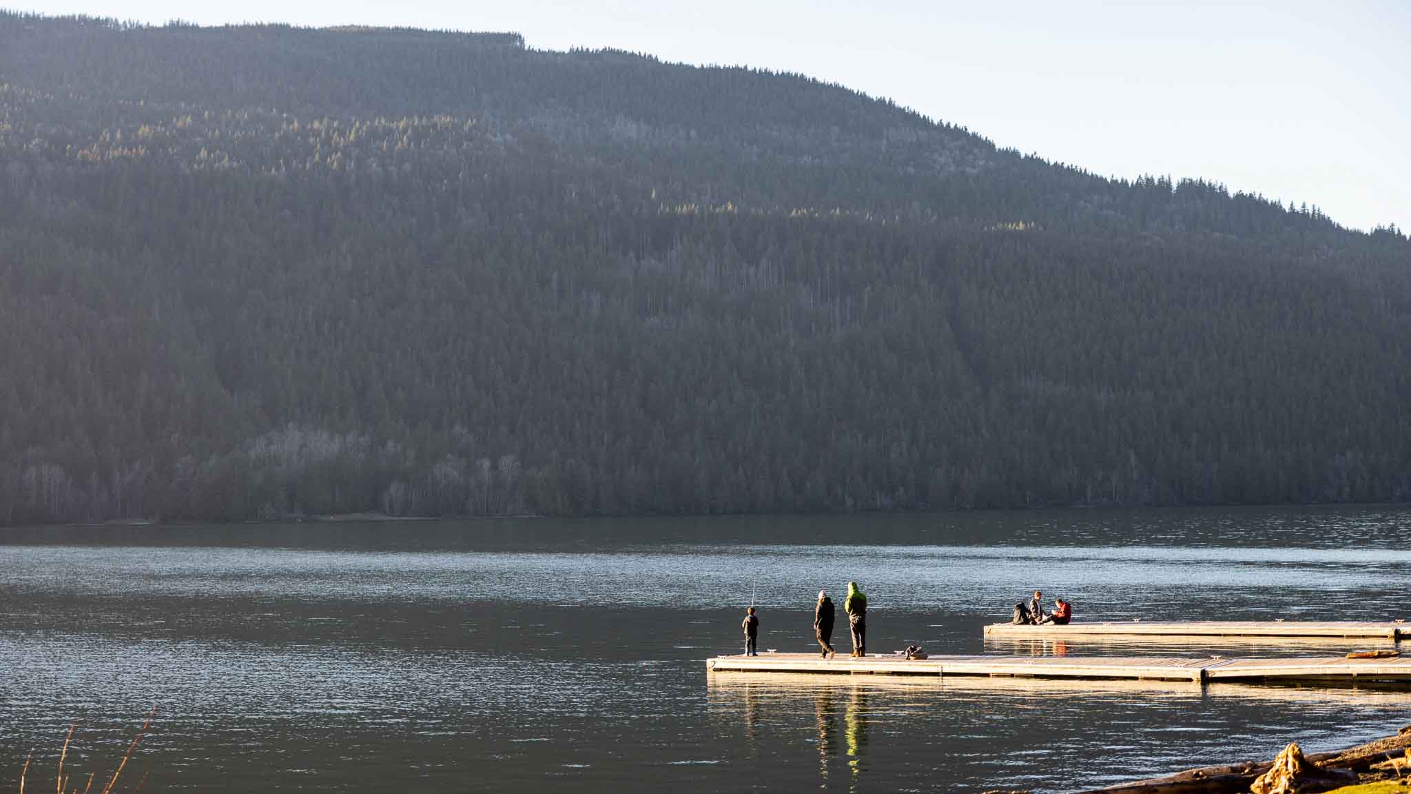 A sunny view around where the water quality testing was happening in Chilliwack. A mountain rises in the background, while in the foreground people stand and sit on two wooden docks which extend into the water - on one, three people are fishing with rods.